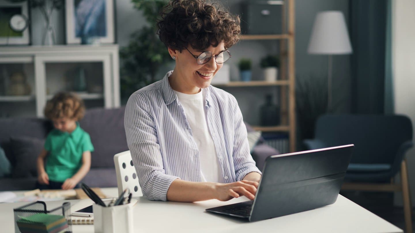 Woman smiles while typing on laptop at desk with child in background.
